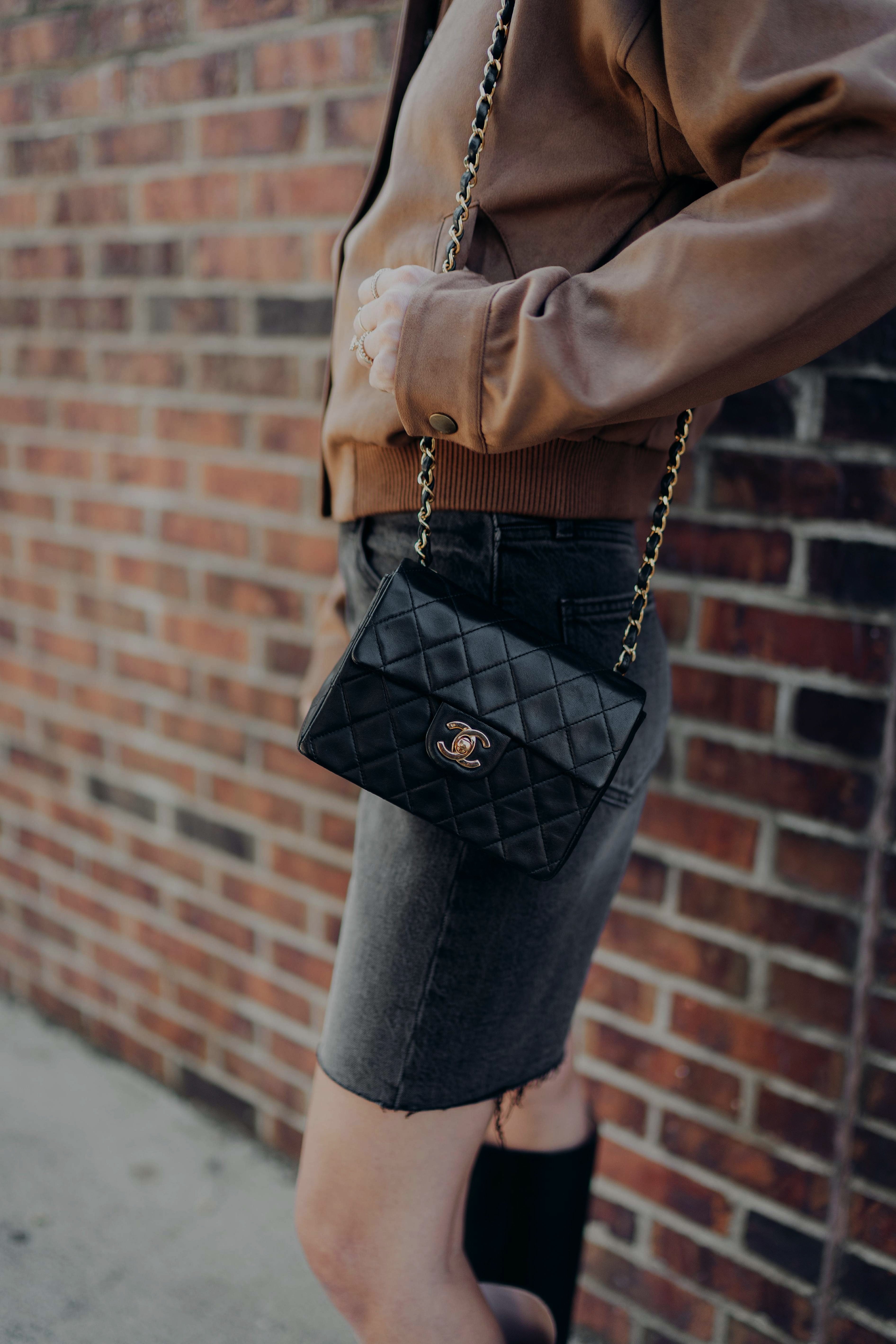 Person holding a Chanel black mini classic flap against a brick wall wearing a brown leather jacket and jean shorts with black boots