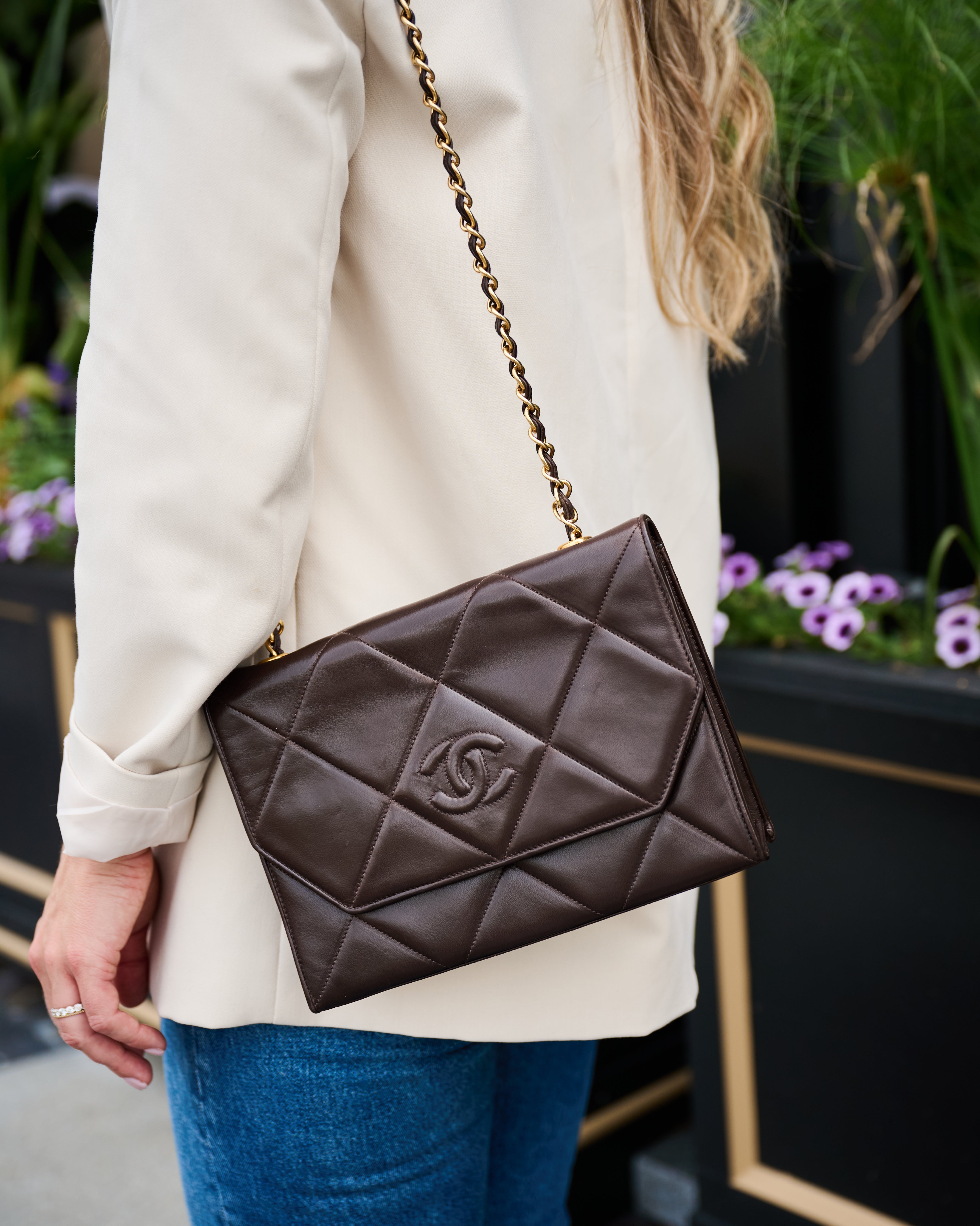 Chanel brown quilted handbag with gold chain strap held by a person wearing a beige coat and blue jeans